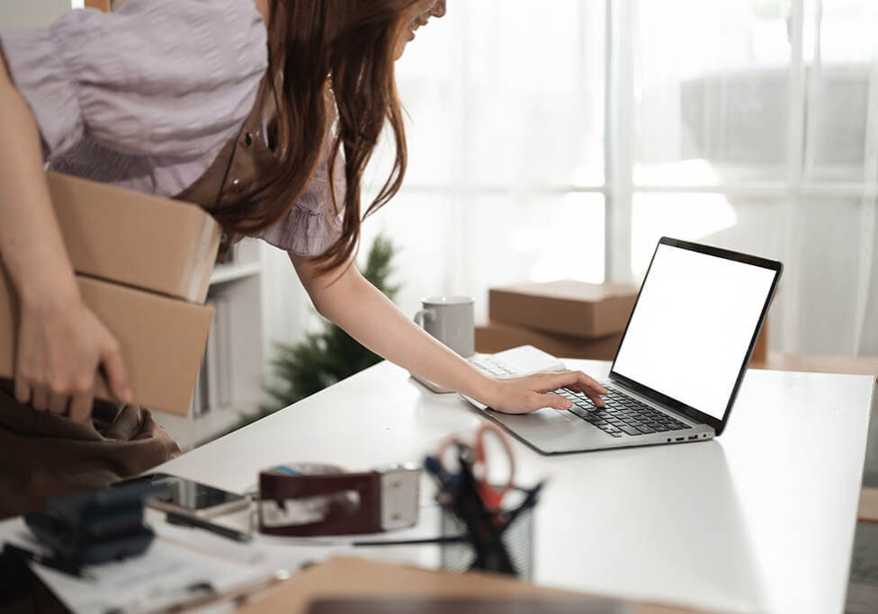 A Close-up of a small business owner multitasking, balancing package shipments while checking orders on a laptop, Empty screen of laptop.